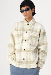 Creme plaid fleecejakke med orange og grå linjer, der har krave, to frontlommer og sorte knapper. Teksturen ser blød og lækker ud.