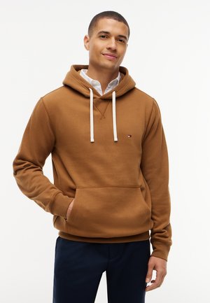 Jonge man draagt een bruine hoodie over een wit shirt, met één hand in de voorzak, staand tegen een effen witte achtergrond.