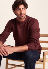 Burgundy knitted sweater with a rounded neckline, ribbed cuffs, and textured fabric. Model seated on a wooden chair, wearing dark jeans.