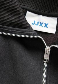 Chaqueta negra con cremallera y cuello de canalé, de tela lisa, cremallera plateada y etiqueta blanca con texto azul "JJXX."