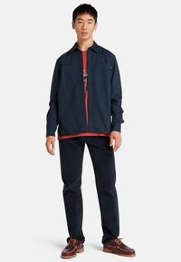 Marine knoopjesjack over een rode graphic tee, gecombineerd met donkere broek en bruine schoenen. Kenmerken omvatten patchzakken en contrasterende texturen.