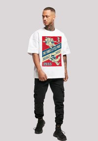 Hvit oversized t-skjorte med et fargerikt grafisk motiv som viser "De tre små grisene" og "Den store stygge ulven", svarte cargo-bukser og joggesko.