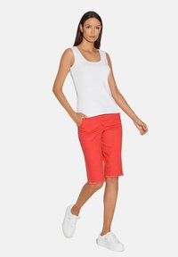 Rode shorts met een rafelige zoom, gecombineerd met een witte mouwloze top. Model draagt witte sneakers en staat met één hand in de zak.