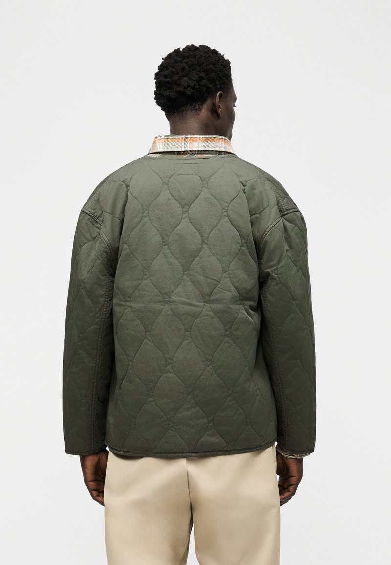 Homme portant une veste matelassée vert olive sur un pantalon beige, vu de dos, sur un fond clair uni.