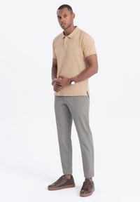 Beige polo tričko, vzorované šedé kalhoty a hnědé tenisky. Má krátké rukávy, límec a decentní logo na tričku.