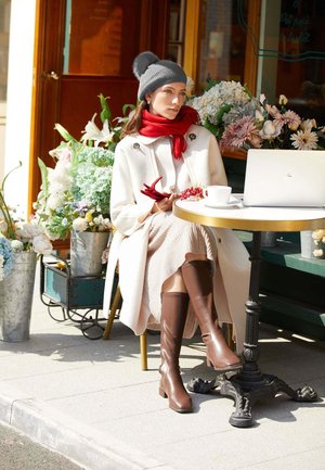 Bottes en cuir marron longues, manteau oversized crème, écharpe rouge, bonnet gris, assise à une table de café avec des fleurs et un ordinateur portable à proximité.