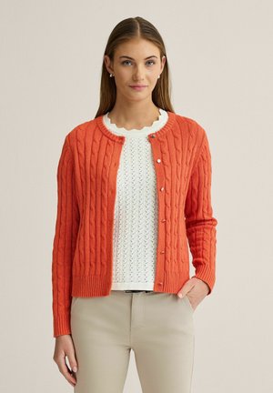 Kvinde iført en orange kabelstrikket cardigan over en hvid strikket top med bølget halsudskæring og lyse beige bukser, stående mod en ensfarvet baggrund.
