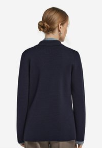 Dunkelblazer aus Navyblau mit langen Ärmeln, subtiler Textur, Kragen und geradem Schnitt. Die Rückansicht zeigt ein einfaches, unaufdringliches Design.