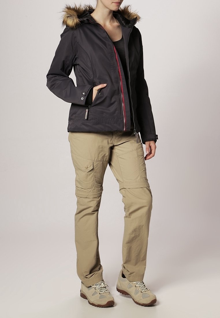 Femme portant une veste d'hiver noire à capuche avec bordure en fourrure, un pantalon cargo beige et des chaussures de randonnée beiges, debout avec une main dans la poche de la veste.