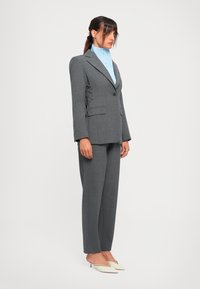 Woman in a gray tailored suit with side lace-up details, light blue high-neck shirt, and white pointed-toe heels standing sideways on white background.
