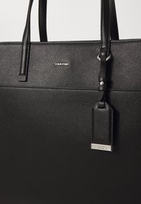 Sac fourre-tout en cuir noir texturé avec double poignée, logo Calvin Klein argenté sur le devant, et une étiquette en cuir suspendue avec détail en métal.