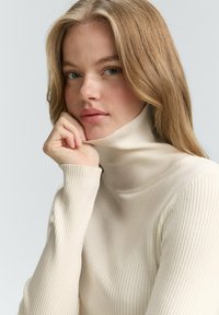 Top bej deschis cu guler înalt, cu textura moale, având mâneci lungi și un guler înalt. Modelul își sprijină bărbia în palmă.