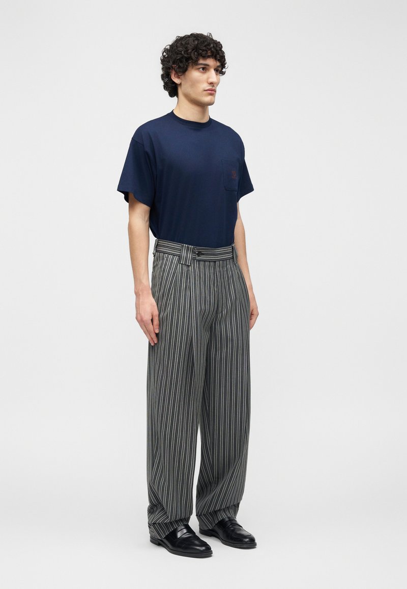 Giovane uomo con capelli ricci che indossa una maglietta blu navy e pantaloni a gamba larga grigi a righe sottili, a vita alta, con scarpe nere.