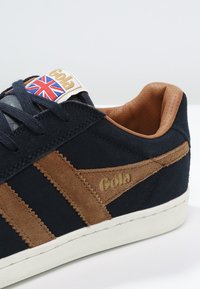 Sneakers Gola in suede blu navy con dettagli marroni, suola in gomma bianca, caratterizzati da un logo Union Jack e una linguetta testurizzata.