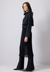 Black two-piece outfit featuring a cropped jacket with a collared neckline and high-waisted pleated pants. Smooth texture, total monochrome design.