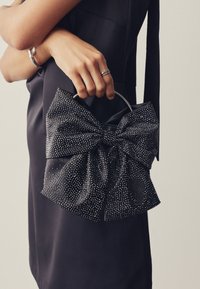 Juodos odos rankinė su dideliu dekoratyviniu kaspinu, padengtu mažais sidabriniais kniedėmis, turinti apvalią rankeną. Dėvima su juoda suknele.