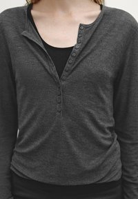Gray long-sleeve shirt with a henley neck, featuring a row of six black buttons. Worn over a black inner layer. Soft texture, casual fit.