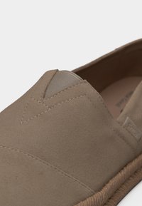 Gros plan sur une chaussure à enfiler en suède taupe avec une insert en élastique en forme de V et une couture détaillée le long de la partie supérieure et du bord de la semelle tissée.
