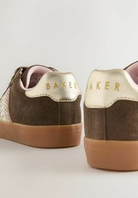 Hnědé semišové tenisky s metalickým zlatým akcentem na patě, na kterých je zlatě embosované slovo "BAKER". Gumová podrážka a růžové podšívání.