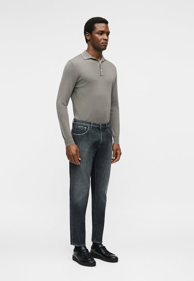 Άνδρας που στέκεται φορώντας γκρι πόλο με μακριά μανίκια, ξεθωριασμένο μαύρο τζιν και μαύρα αθλητικά παπούτσια, σε απλό ανοιχτό φόντο.