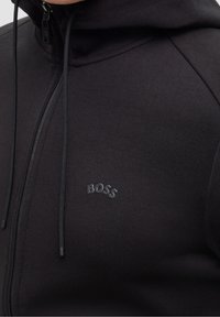 Närbild av en svart zip-up hoodie med snoddar och en liten upphöjd "BOSS"-logotyp på bröstet.