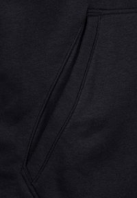 Detailní záběr na černou látkovou kapsu s zesíleným švem, pravděpodobně součást oblečení jako jsou tepláky nebo mikina.