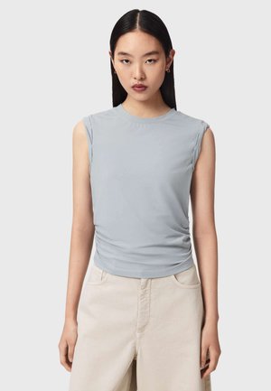 Junge Frau mit langen schwarzen Haaren, die ein hellblaues ärmelloses Oberteil und beige Hose trägt und vor einem schlichten grauen Hintergrund steht.