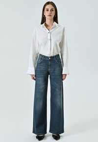 Witte button-up shirt met lange mouwen en gouden knoopdetails, gecombineerd met blauwe wijde spijkerbroek en zwarte puntige schoenen.