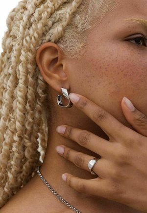 Zilveren oorring met een golvend ontwerp, voorzien van een gepolijste afwerking. De hand van het model raakt het oor aan, waarbij de zachte huid en de ring zichtbaar zijn.