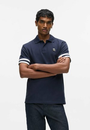 Jonge man met een marineblauw poloshirt met witte strepen op de mouwen en blauwe spijkerbroek, staand met gekruiste armen tegen een effen witte achtergrond.