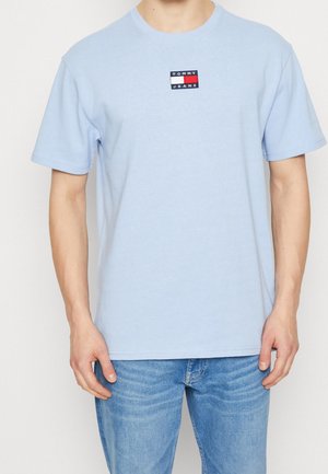 T-shirt en coton bleu clair avec des manches courtes. Présente un petit logo rectangulaire en rouge, blanc et bleu sur la poitrine.