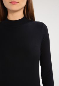 Schwarzes Langarmoberteil aus geripptem Material, mit hohem Kragen und tailliertem Design, das eine glatte Textur und einen subtilen Stretch aufweist.