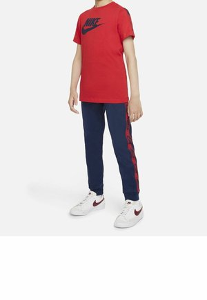 Dziecko ubrane w czerwoną koszulkę Nike, granatowe spodnie dresowe Nike z czerwonymi bocznymi paskami oraz białe sneakersy Nike z bordowymi logo, stojące.