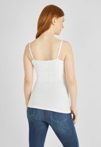 Fehér pamut tank top, állítható pántokkal és egyszerű, testhezálló kialakítással. Kék farmernadrággal viselve.