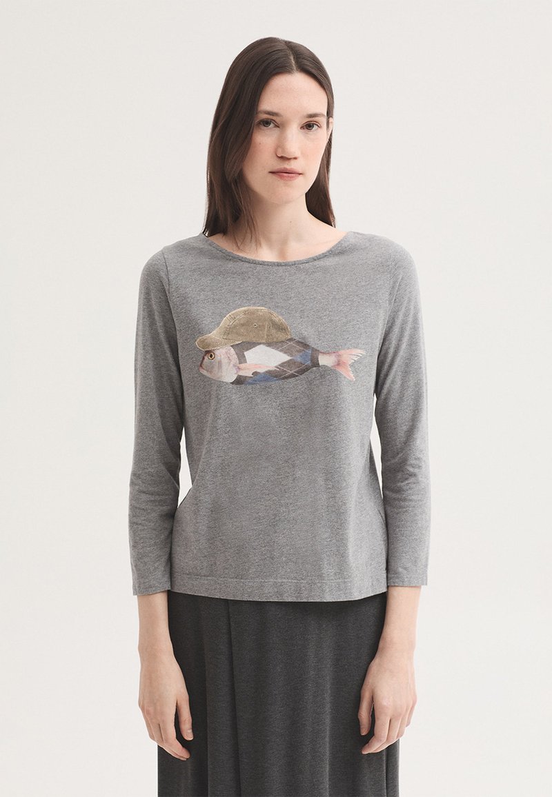 Jeune femme aux cheveux bruns raides porte un t-shirt gris à manches longues avec un motif de poisson portant un chapeau sur le devant.