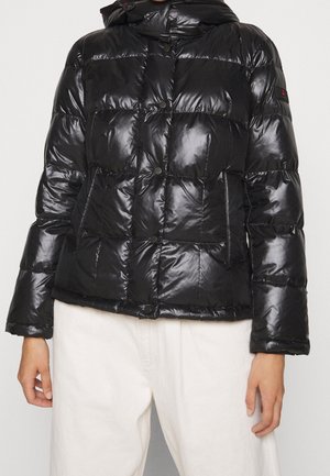 Osoba ubrana w czarną błyszczącą pikowaną kurtkę z zatrzaskami i bocznymi kieszeniami, w połączeniu z białymi spodniami na jednolitym tle.