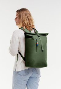Mochila verde de tipo roll-top con correas ajustables, diseño elegante, bolsillo con cremallera, textura minimalista y borlas de color como acento.