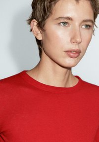 Roter Strickpullover mit rundem Halsausschnitt, der eine glatte Textur und eine figurbetonte Passform aufweist. Der Kopf und die Schulter des Modells sind teilweise sichtbar.