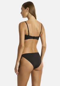 Modelka w czarnym bikini, widziana od tyłu, prezentująca cienkie ramiączka i niskie majtki, na jednolitym białym tle.