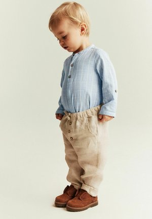 Tout-petit portant une chemise bleu clair boutonnée, un pantalon beige et des chaussures marron, debout et regardant vers le bas sur un fond uni.