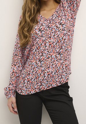 Kobieta w bluzce z długim rękawem w różowy kwiatowy wzór, w połączeniu z czarnymi spodniami, stojąca na jednolitym tle.