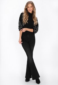 Pulover negru cu broderie florală albă pe mâneci, asortat cu blugi negri evazați și cizme negre până la gleznă. Textură netedă, design ajustat.