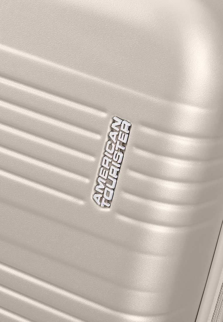 Valise rigide beige avec texture à côtes verticales. Présente un logo argenté "American Tourister" embossé sur la surface.