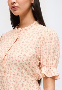 Γυναίκα με ανοιχτό ροζ μπλούζα με μικρό floral print, βολάν στο γιακά και φουσκωτά κοντά μανίκια με ελαστικές μανσέτες.