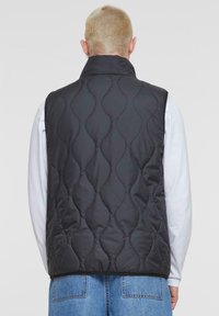 Schwarzige gesteppte Weste mit hohem Kragen, mit einem gewellten Muster, getragen über einem weißen Langarmshirt, kombiniert mit blauen Jeans.