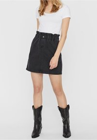 Minifalda de mezclilla negra con cintura fruncida, cierre frontal de botones y bolsillos laterales, combinada con una camiseta blanca ajustada y botas negras.