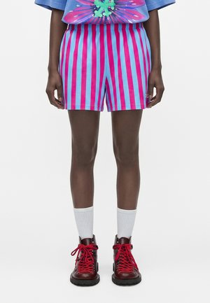 Persona con pantalones cortos de rayas verticales azules y rosas, calcetines blancos hasta la mitad de la pantorrilla y botas rojo oscuro con cordones rojos, de pie sobre un fondo blanco.