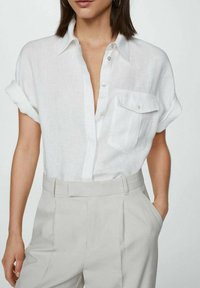 Camicia bianca in lino con maniche corte arrotolate e taschino sul petto, abbinata a pantaloni alti grigi chiari.