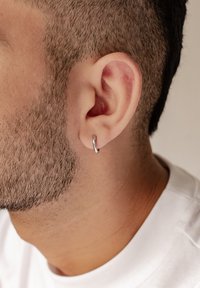 Orecchino a cerchio in argento attaccato a una barba regolare con leggera peluria. L'orecchino ha una finitura lucida che riflette la luce in modo sottile.