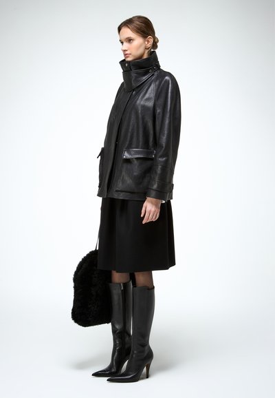 Mujer con chaqueta de cuero negra, falda hasta la rodilla y botas altas negras sosteniendo un bolso de piel negra, posando contra un fondo claro.